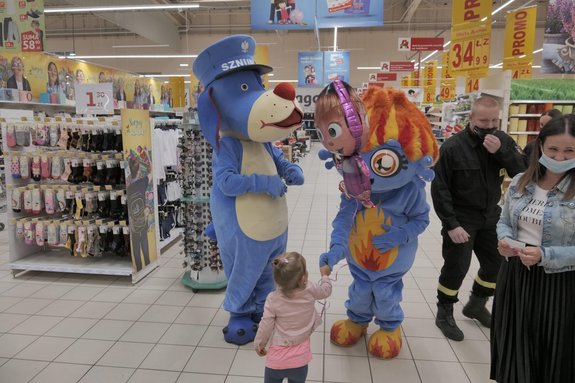 Zdjęcie kolorowe. Widoczne maskotki Sznupka i Pożorka, które chodzą na terenie sklepu w asyście policjantów i klientów