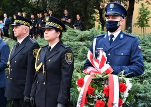 Kolorowe zdjęcie, na którym jest umundurowany policjant trzymający wieniec kwiatów. Obok niego stoją umundurowani funkcjonariusze straży miejskiej.