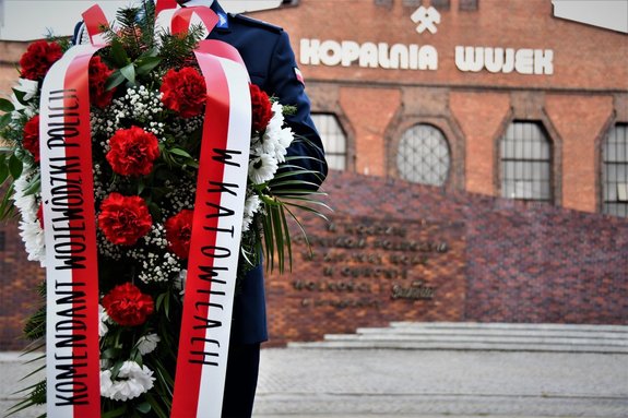 Kolorowe zdjęcie przedstawia wieniec biało-czerownych kwiatów, które trzyma policjant. Widoczny tylko jego korpus, w tle Kopalnia Wujek.