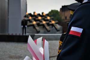 Kolorowe zdjęcie. Na pierwszym planie flaga Polski na ramieniu policjanta, w tle palące się na krzyżach przed kopalnią znicze, które upamiętniają poległych górników.