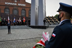 Kolorowe zdjęcie. Na pierwszym planie policjant trzymający wieniec, w tle palące się na krzyżach przed kopalnią znicze, które upamiętniają poległych górników oraz Kopalnia Wujek.