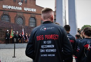 Kolorowe zdjęcie. Na pierwszym planie uczestnik biegu, widoczny napis na plecach jego bluzy - Bieg Pamięci, ku czci męczenników stanu wojennego Katowice-Warszawa, w tle kopalnia Wujek.