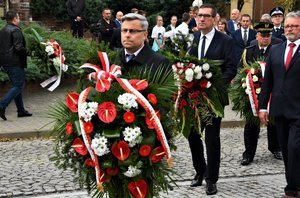 Kolorowe zdjęcie. Wojewoda Śląski trzyma wieniec kwiatów. Za nim inne osoby, które będą składać kwiaty pod pomnikiem.