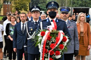 Kolorowa fotografia. Policjant trzyma wieniec kwiatów, za nim stoją Komendanci Policji oraz pozostali uczestnicy uroczystości.