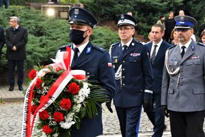 Kolorowa fotografia. Policjant trzyma wieniec kwiatów, za nim stoją Komendanci Policji.