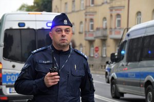 Zdjęcie kolorowe. Widoczny umundurowany policjanci, w tle radiowóz