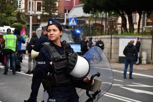 Zdjęcie kolorowe. Umundurowana policjantka idąca jezdnią. W tle policyjne radiowozy oraz tłum ludzi.