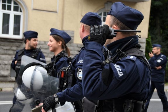 Zdjęcie kolorowe. Pięciu umundurowanych policjantów. Policjant znajdujący się na pierwszym planie trzyma w ręce radiostację.