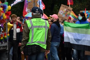 Zdjęcie kolorowe. Na pierwszym planie nieumundurowany policjant w kamizelce odblaskowej z napisem &quot;Zespół Antykonfliktowy Policja&quot;. W tle tłum ludzi biorących udział w marszu.