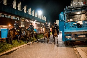 Policjanci policji konnej na koniach, podczas zabezpieczenia meczu wieczorową porą. Z prawej strony fragment armaty wodnej.