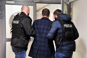 Policjanci w czarnych kamizelkach z napisem Policja prowadzą zatrzymanego mężczyznę korytarzem.