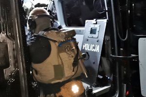 Na zdjęciu policyjny komandos w pełnym umundurowaniu z młotem na plecach, obok niego tarcza.