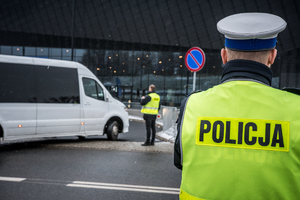 Umudurowany fukcjonariusz policji,w tle funkcjonariusz Służby Ochrony Państwa kontrolujacy wjeżdżający autokar