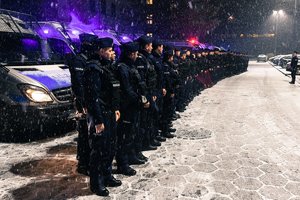 Stojący w szeregu umundurowani funkcjonariusze policji, w tle radiowozy policyjne z włączonymi sygnałami świetlnymi pojazdu uprzywilejowanego.