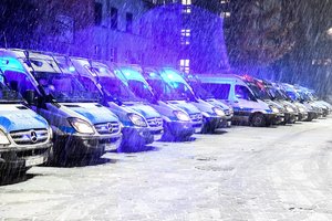 Rząd radiowozów policyjnych stojących przed budynkiem koszarowym oddziału prewencji z włączonymi sygnałami świetlnymi pojazdu uprzywilejowanego.