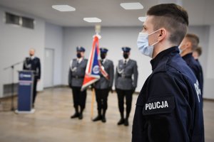 Na pierwszym planie umundurowany, nowo przyjęty policjant, w tle poczet sztandarowy oraz lektor prowadzący uroczystość ślubowania.