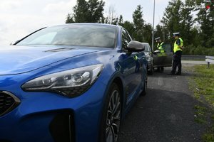 na pierwszym planie nieoznakowany radiowóz kia stinger. W tle policjanci ruchu drogowego podczas kontroli drogowej.