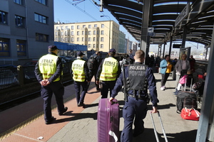 w centralnej części zdjęcia policjant z walizkami idący peronem dworca, po prawej stronie funkcjonariusze straży ochrony kolei