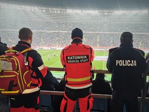 Zdjęcie kolorowe. Widoczny policjant i ratownicy na stadionie