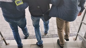 Widoczna osoba prowadzona przez dwóĽch policjantów, która ma założone kajdanki zespolone