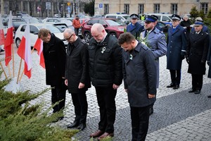 Osoby oddają hołd przed pomnikiem, za nimi stoi delegacja służb mundurowych, która nadchodzi z kwiatami.