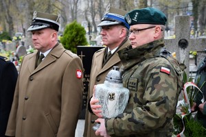 Goście biorący udział w uroczystości na cmentarzu. Jeden z mundurowych trzyma zapalony znicz.