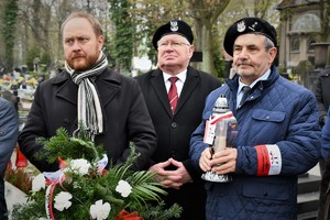 Goście biorący udział w uroczystości na cmentarzu. Jeden z mężczyzn trzyma zapalony znicz.