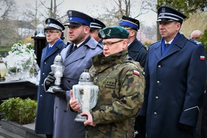 Delegacja służb mundurowych stoi przed grobem. Dwóch mundurowych trzyma zapalony znicz.