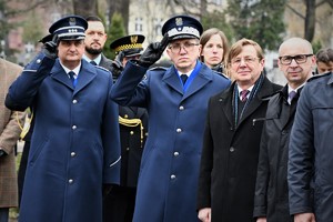 Goście biorący udział w uroczystości stoją na cmentarzu. Wśród nich Komendant Wojewódzki Policji w Katowicach i Komendant Szkoły Policji w Katowicach. Obaj policjanci oddają honor.