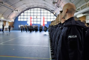 Nowi policjanci stoją w szeregu, w tle biało-czerwona flaga.