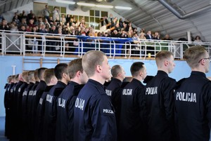 Nowo przyjęci policjanci stoją w szeregu, w tle widoczny ich bliscy, którzy wzięli udział w uroczystości.