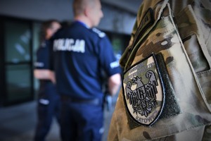 Ramie policjanta z naszywką pododdział kontrterrorystyczny. W tle umundurowani policjanci.