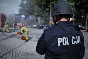 Na zdjęciu umundurowany policjant. Widoczne jego plecy i napis policja, w tle funkcjonariusze straży pożarnej.