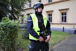 Na zdjęciu umundurowany policjant w hełmie i kamizelce odblaskowej. W rękach trzyma pistolet maszynowy.