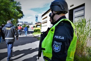 Na tle kopalni widać policjantów biorących udział w ćwiczeniach, na pierwszym planie z prawej strony widać ustawionego bokiem umundurowanego policjanta w kasku taktycznym oraz kamizelce  odblaskowej, po lewej stronie widać idących policjantów ubranych po cywilu. W oddali zdjęcia po lewej stronie widać ustawiony tyłem żółty autobus.