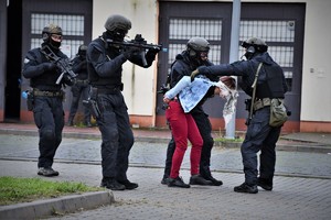 Na zdjęciu umundurowani policjanci z bronia maszynową prowadzą osobę