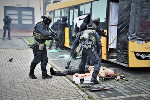 Zdjęcie. Widoczni umundurowani policjanci z bronią maszynową przed autobusem, oraz osoba leżąca na chodniku