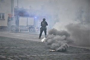Zdjęcie. Widoczny umundurowany policjant, granaty dymne, w tle wóz strazy pożarnej