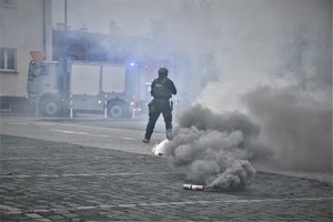 Zdjęcie. Widoczny umundurowany policjant, granaty dymne, w tle wóz strazy pożarnej