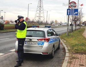 na zdjęciu policjant z ruchu drogowego ubrany w kamizelkę odblaskową koloru żółtego trzyma w ręce radar i mierzy prędkość pojazdom w rejonie przejścia dla pieszych, w tle widać radiowóz