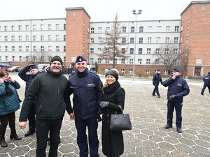 Na zdjęciu widzimy uśmiechniętego policjanta zaraz po ślubowaniu , który stoi pomiędzy swoimi rodzicami.