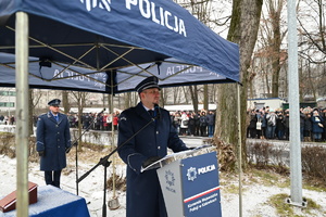 Na zdjęciu widoczny policjant prowadzący uroczystość, natomiast w tle widać rodziny nowoprzyjętych policjantów.