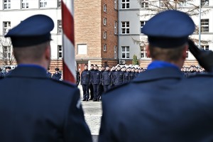 zdjęcie przedstawia dwóch policjantów stojących tyłem w tle budynek, a przed nim policjanci w szeregach