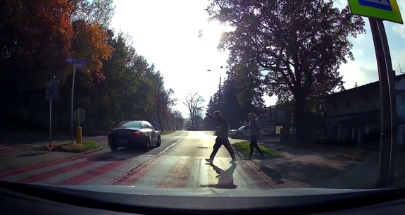 Widok z kamery samochodowej. Na zdjęciu przejście dla pieszych, na które z prawej strony wchodzi dwóch mężczyzn. Z lewej strony, za przejściem samochód mercedes.
