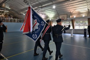 Zdjęcie. Widoczny policyjny poczet sztandarowy na sali sportowej. Na dalszym planie goście uroczystości
