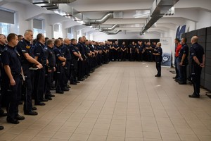 Zdjęcie. Widoczna sala, na której w szeregu stoi grupa kilkudziesięciu umundurowanych policjantów