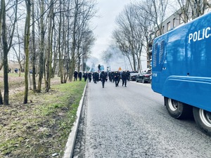 Umundurowani policjanci idący ulicą, a na pierwszym planie jadący policyjny pojazd