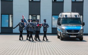Sześciu policjantów kompanii honorowej idzie ze złożoną flagą  i sztandarem, obok radiowóz
