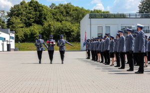 Trzech policjantów kompanii honorowej ze złożona flagą idzie wzdłuż ustawionych na placu mianowanych