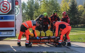 Zielone drzewa na tle płyty lądowiska, dwóch ratowników medycznych przygotowuje nosze z poszkodowanym do umieszczenia we wnętrzu ambulansu Pogotowia Ratunkowego. Medykom asystuje trzech TOPR-owców i policyjny lotnik.